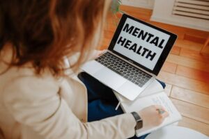 A woman participating in an online therapy session, taking notes with a laptop displaying 'Mental Health'.